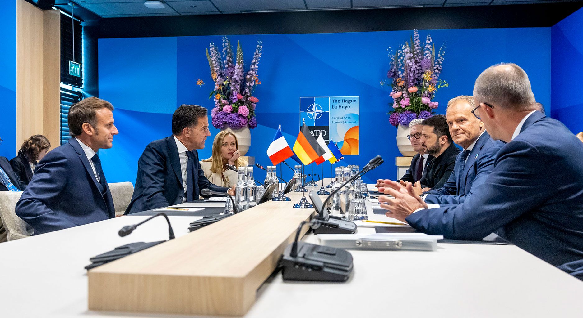 Auf beiden Seiten eines Konferenztisches sitzen fünf Staats- und Regierungschefs europäischer Staaten, der NATO Generalsekretär und der ukrainische Präsident. Zwischen Ihnen stehen Flaggen der jeweiligen Länder, im Hintergrund hängt das Poster des NATO Gipfels 2025