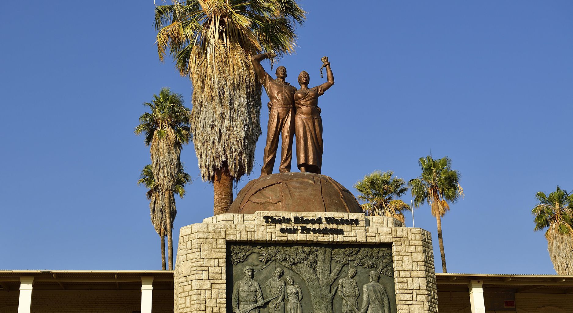The picture shows the Genocide Memorial in Windhoek, Namibia which commemorates the extermination order against Ovaherero and Nama following their rising aganist German rule in Namibia. The Genocide memorial is made of stone with a plaque showing people being hanged and german officers with guns standing in front of them. Atop the memorial two bronze figures raise their fists. On the stone it reads “Their Blood Waters our Freedom”