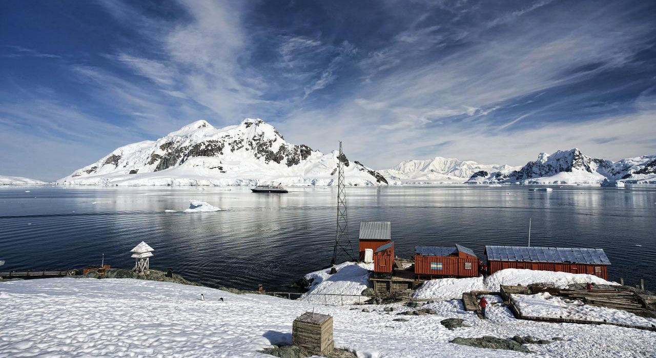 Antarctic coastline with snow mountain range visible in the distance. Three small red buildings are visible in the front right lower corner.
