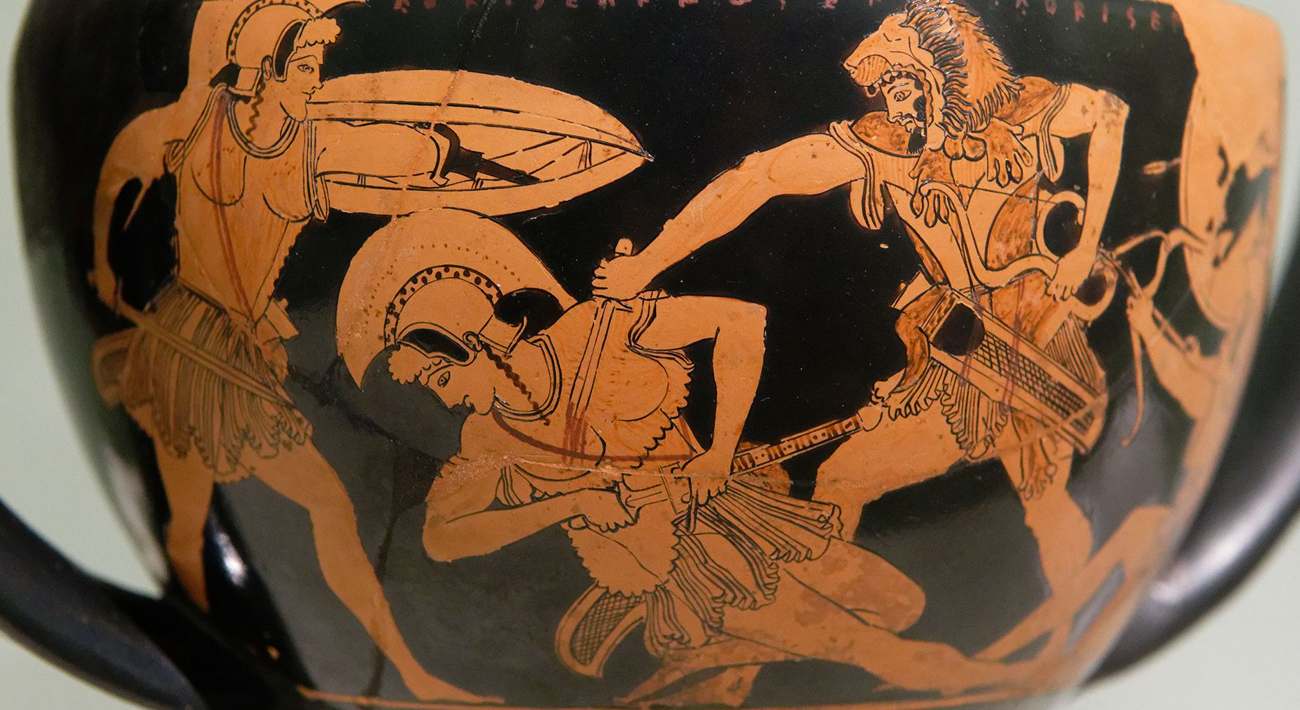 Schwarze antike Vase, auf welcher drei Personen zu sehen sind. Von links nach rechts: Eine Frau mit leichter Rüstung und Helm, welche mit Schild und Schwert ausholt; eine Frau in leichter Rüstung und Helm, welche ihr Schwert zieht; sie fällt zu Boden, weil der Mann rechts sie mit einem Dolch ersticht. Er trägt eine leichte Rüstung und ein Löwenfell. In seiner linken Hand hält er einen Bogen.