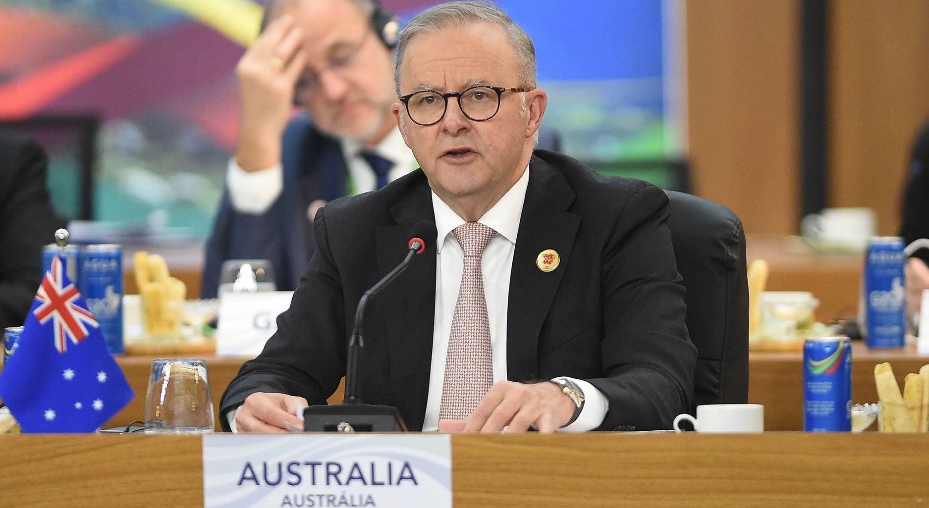 A man in a suit sits at a table with a microfone in front of him. next to him stands a tiny australian flag.