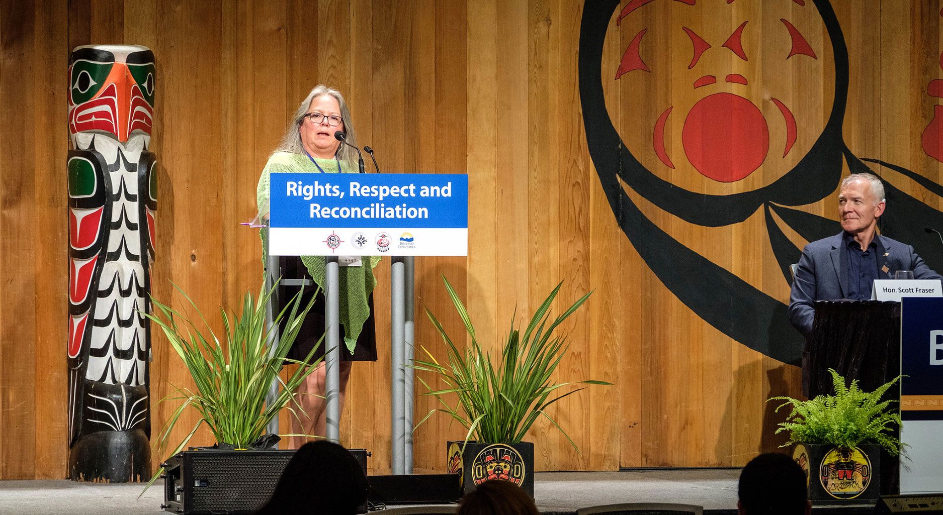 A woman stands on a stage with indigenous insigna behind her