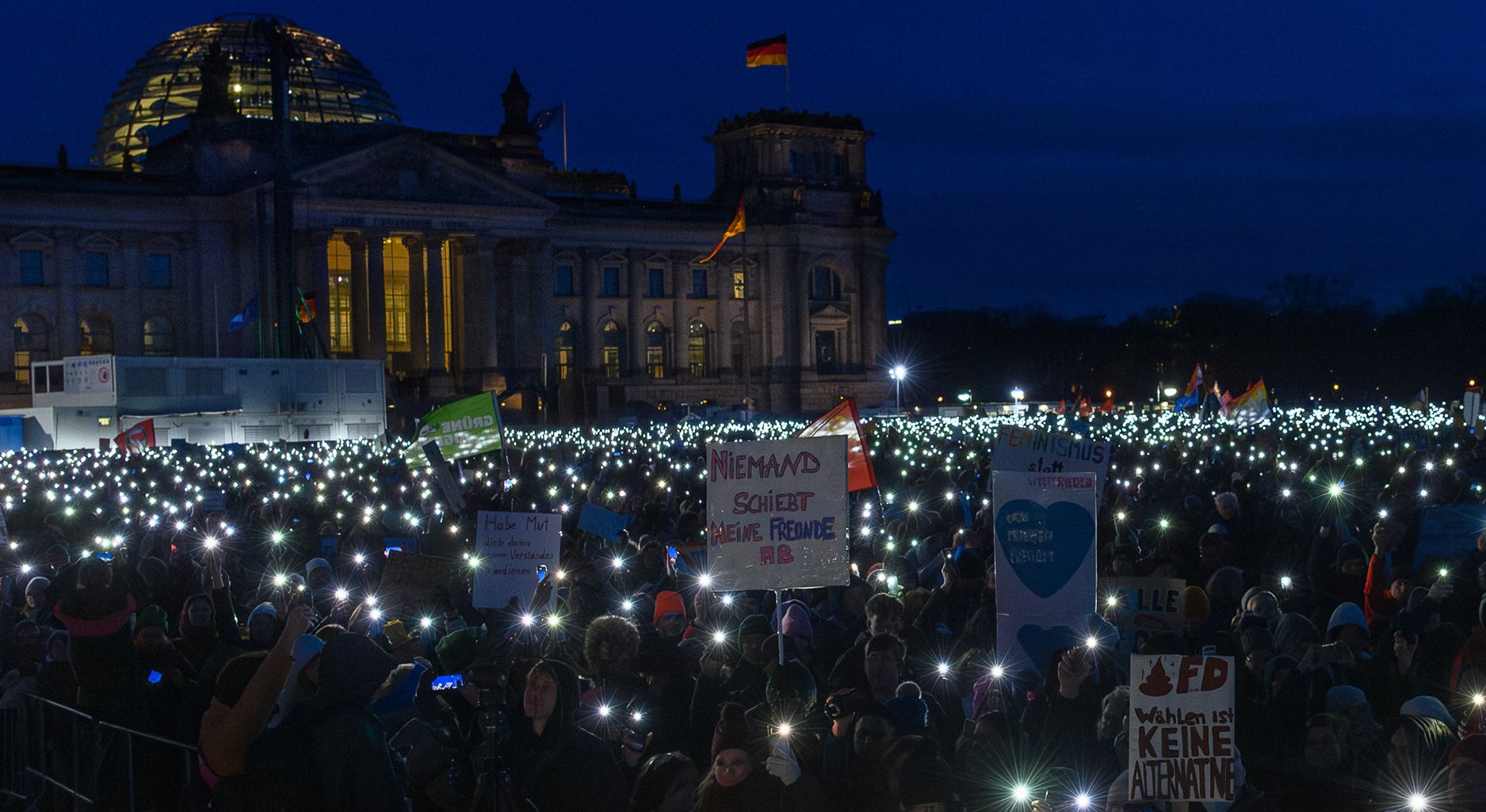 Das Bild zeigt eine große Menschengruppe im dunklen vor dem Bundestag. Sie halten Lichter in die Höhe und Schilder, die Protest gegen die AfD ausdrücken.
