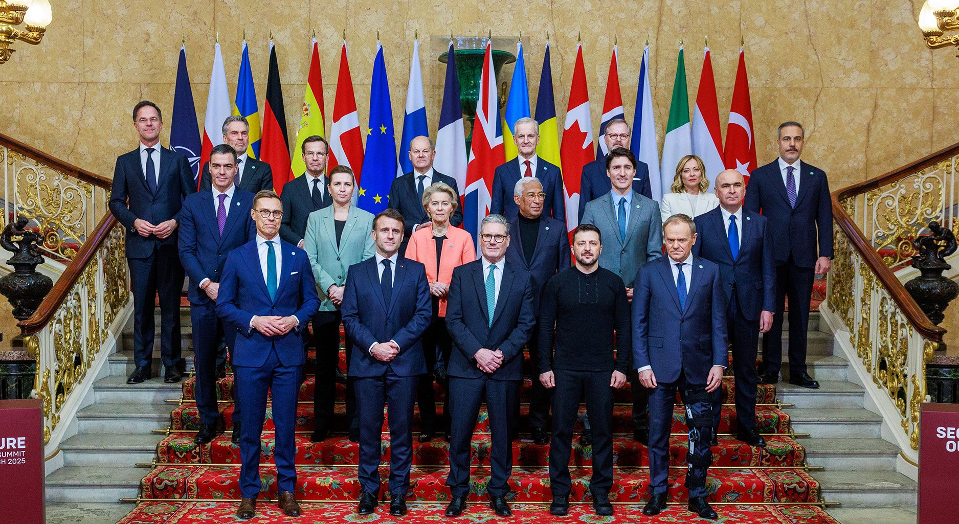 Group photo of formally dressed people on a staircase in front of a row of flags.