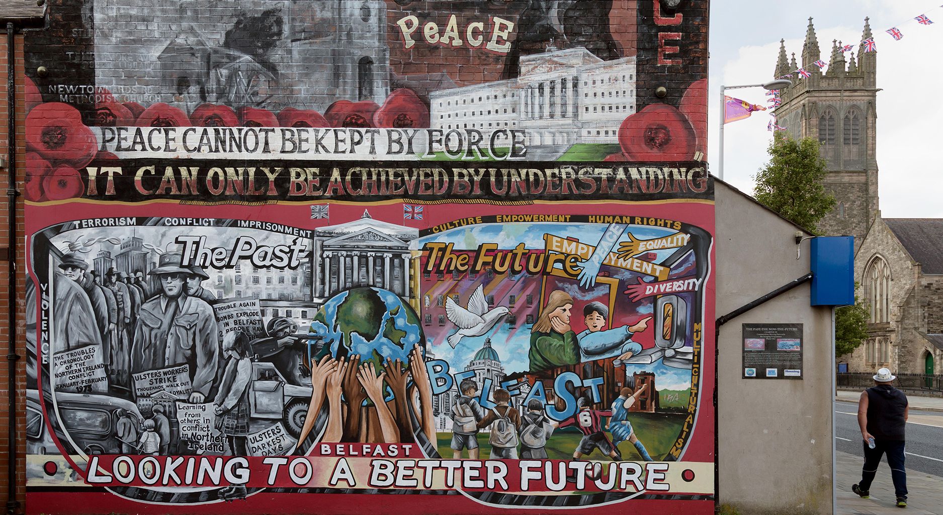 Wandgemälde, auf dem verschiedene Aufschriften zu lesen sind: Peace cannot be kept by force. It can only be achieved by understanding. Darüber sind verschiedene Gebäude und das Wort Peace abgebildet. Auf der unteren Hälfte sind links in Grautönen Menschen in Uniformen abgebildet, daneben Autos und Schilder, deren Beschriftung kaum erkennbar ist. Darüber steht: The Past. Terrorism, Conflict, Imprisonment. In der Mitte ist eine Weltkugel, die von zahlreichen Händen getragen wird. Rechts davon steht: The Future. Daneben sind bunte Arme, die mit Begriffen beschriftet sind, wie: Peace, Equality, Employment, Diversity. Darunter sind Menschen abgebildet, die Fußball spielen und die das Wort Belfast aus Buchstaben vor einem offiziell aussehenden Gebäude aufbauen. Darüber fliegt eine Friedenstaube. Ganz unten steht der Text: Belfast looking to a better future.]