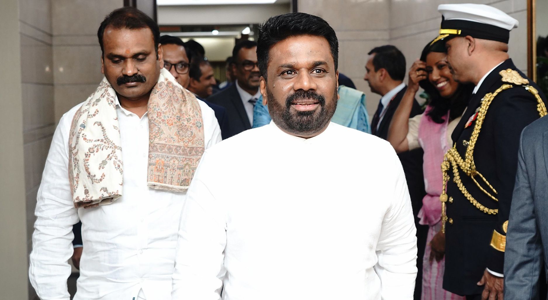 A man formally dressed in white walks towards the camera, followed by other formally dressed people. Others stand on the side including people dressed in military uniform