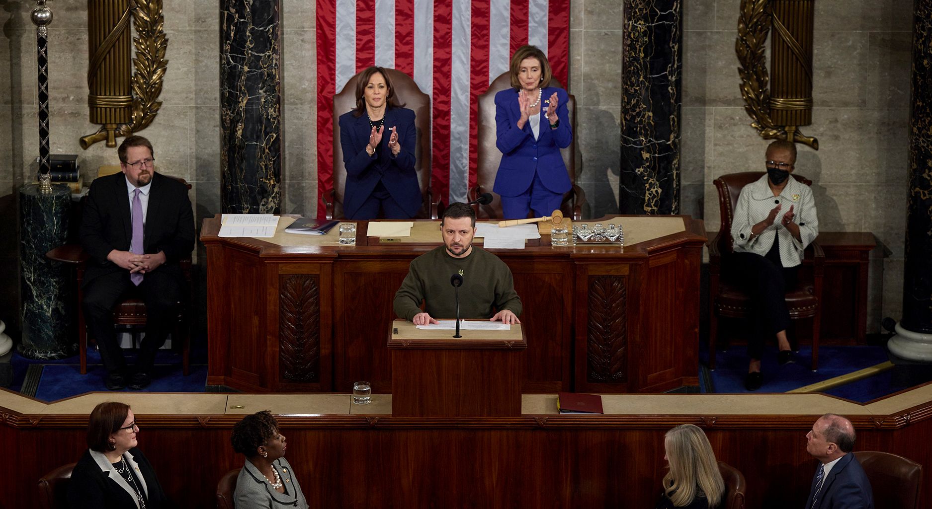 People stand in an old hall on wooden stands and applaud a man who is giving a speech. An american flag in the background on the wall.