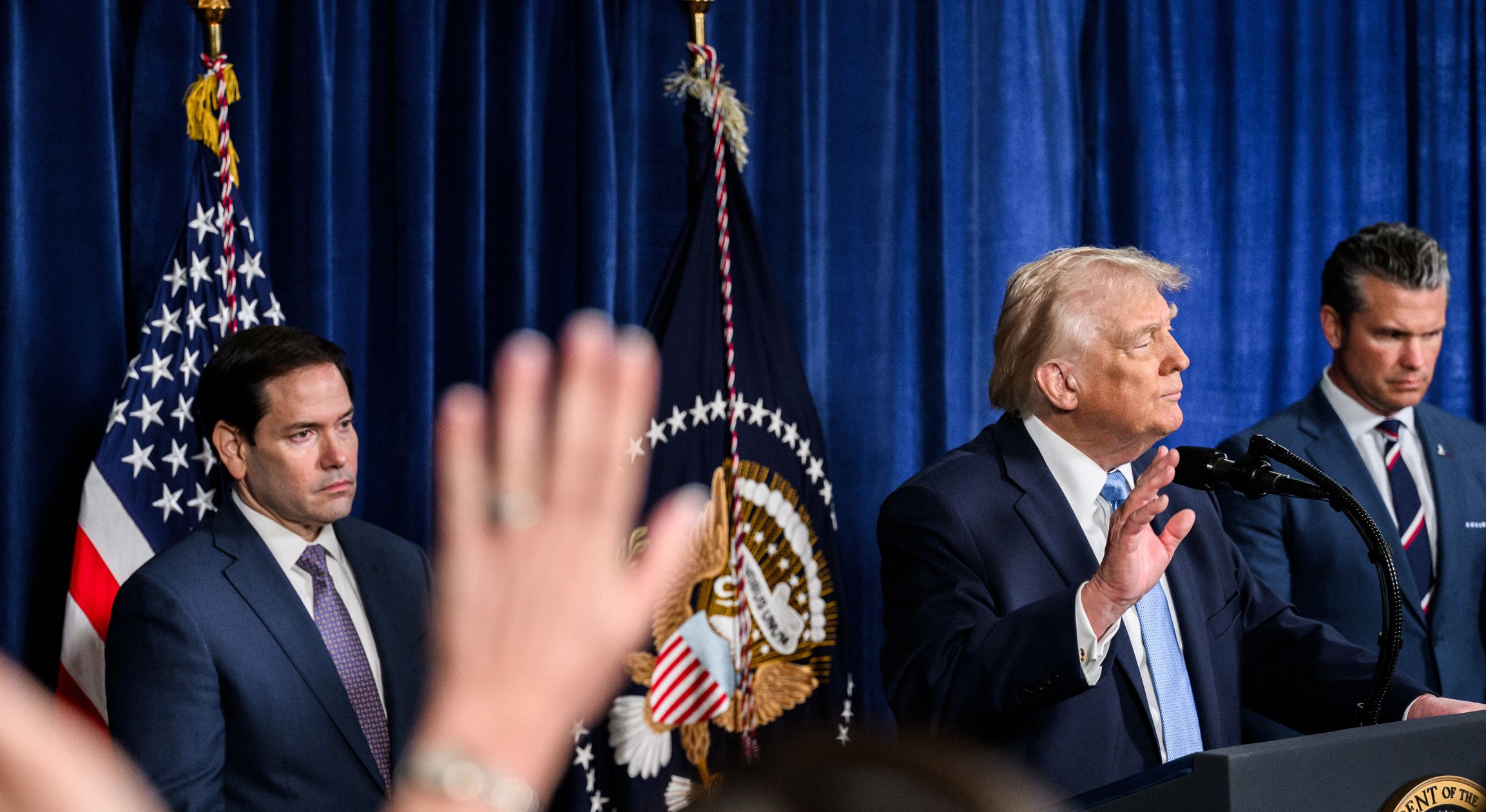 Donald Trump stands at a lectern next to the US flag. Government officials can be seen next to him, with a journalist's raised hand in the foreground.