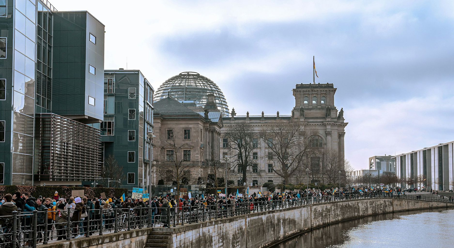 Eine große Gruppe Menschen mit Plakaten, die die Ukraine-Flagge zeigen, laufen links nebem einem Fluss entlang. Im Hintergrund ist das Gebäude des Deutschen Bundestags mit der Glaskuppen zu sehen.
