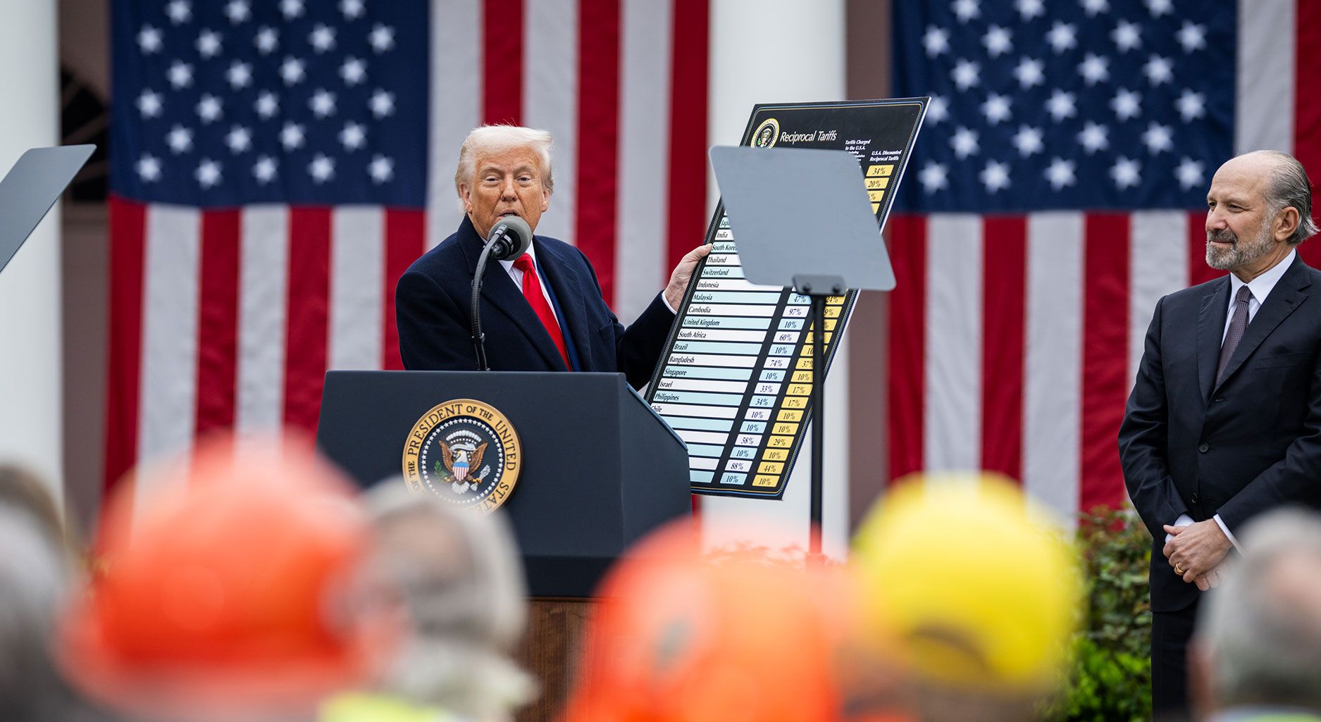 Trump hält eine Karte mit Zöllen in der Hand. Er steht vor zwei riesigen amerikanischen Flaggen.
