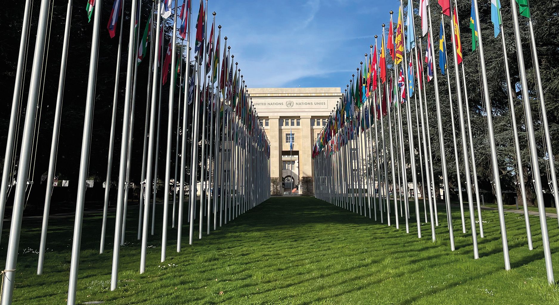 On the left and right side of the picture, national flags stand on grass. The UN building in Geneva stands in the middle of the flags.