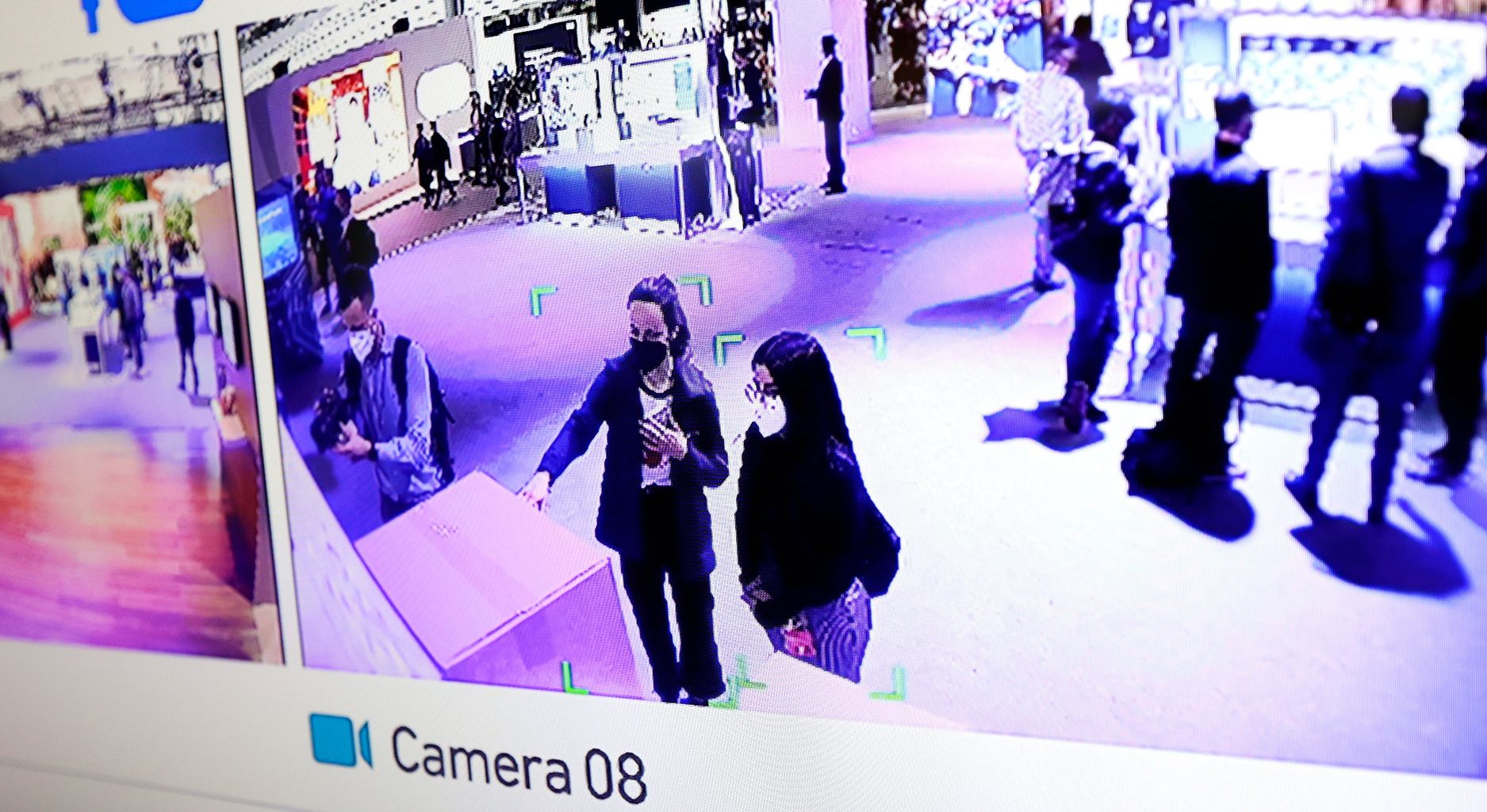 Image shows a screen with footage from two surveillance cameras and the caption "Camera 08" underneath. The footage shows an open room with several people, in the front you can see two women wearing medical masks