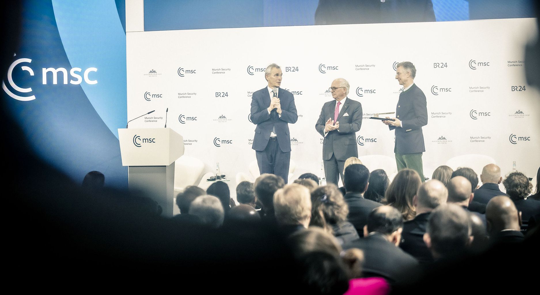 Photo of three men in suits on a stage in front of a crowd of people. To their left is a blue wall with the msc logo.