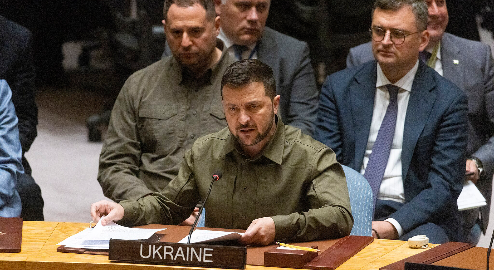 Volodymyr Zelenskyy sitting at a conference desk and speaking into a microphone. In front of him on the desk is a nameplate with "UKRAINE" written on it.