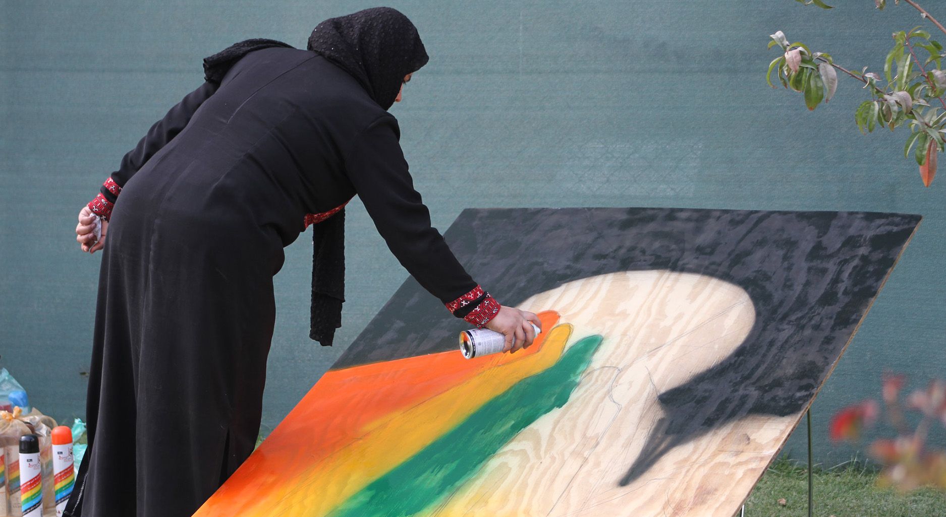 Frau mit Kopftuch und langer Kleidung besprüht eine große Holzplatte mit bunten Farben.