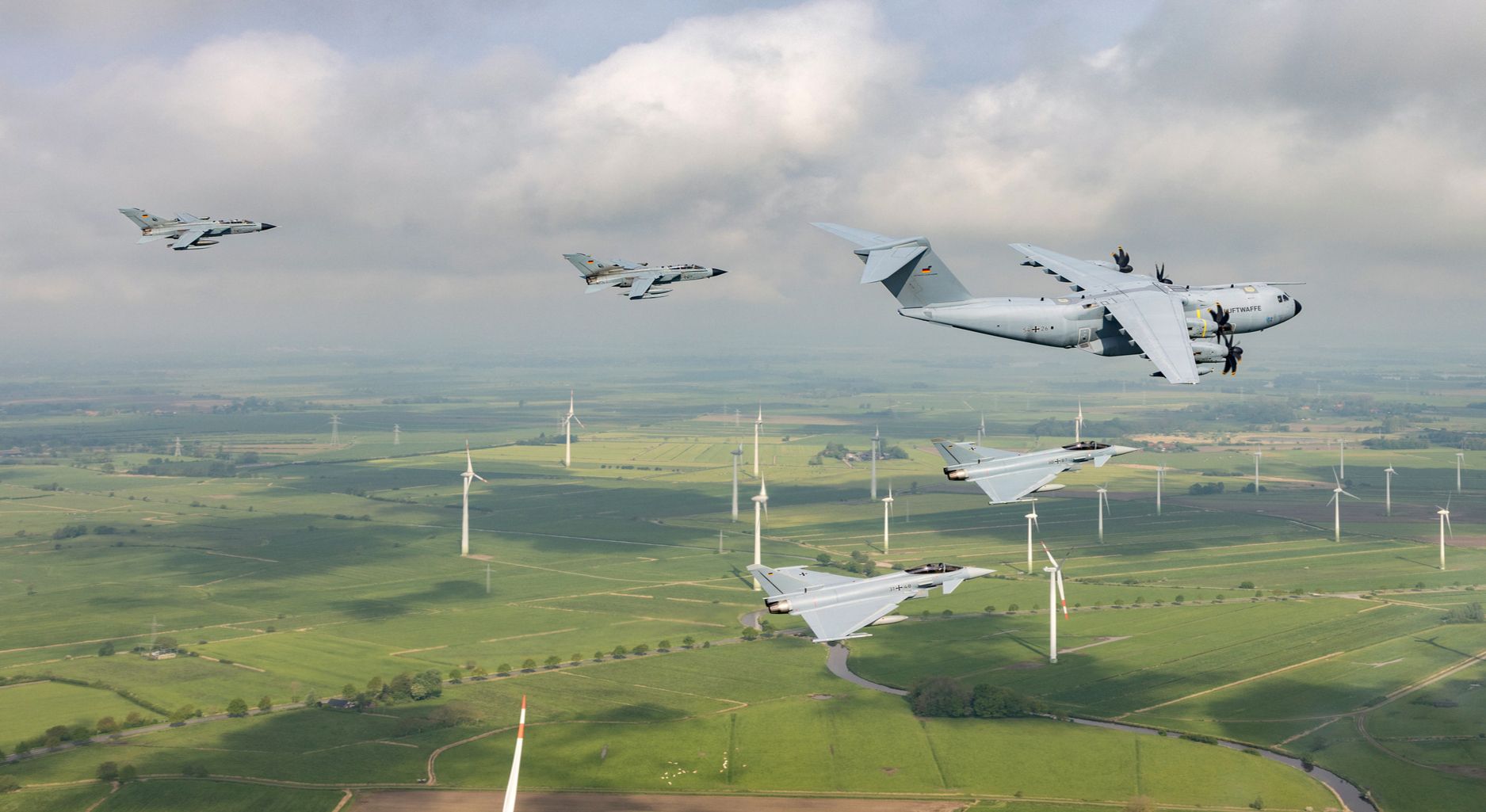 Aerial view; fighter jets flying in the foreground; wind turbines scattered across a grassy landscape in the background