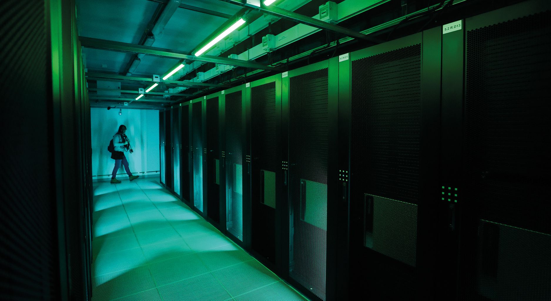 Grünlich beleuchteter Gang eines Serverraums, im Hintergrund geht eine Person vorbei