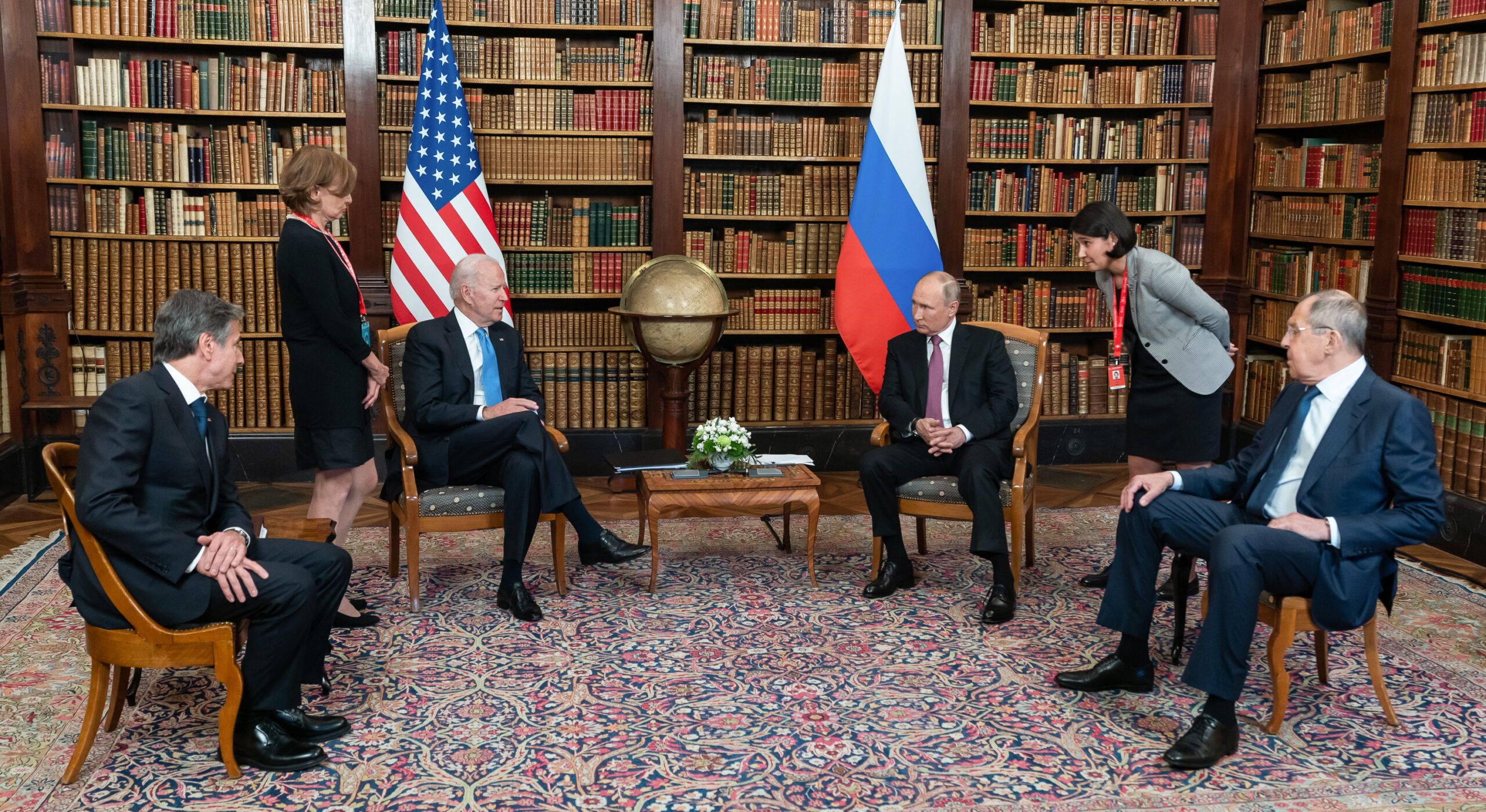 Image shows a meeting between Biden, Putin, Blinken and Lawrow as well as two translators. They are sitting in front of a wall of bookshelves and the US and Russian flags.