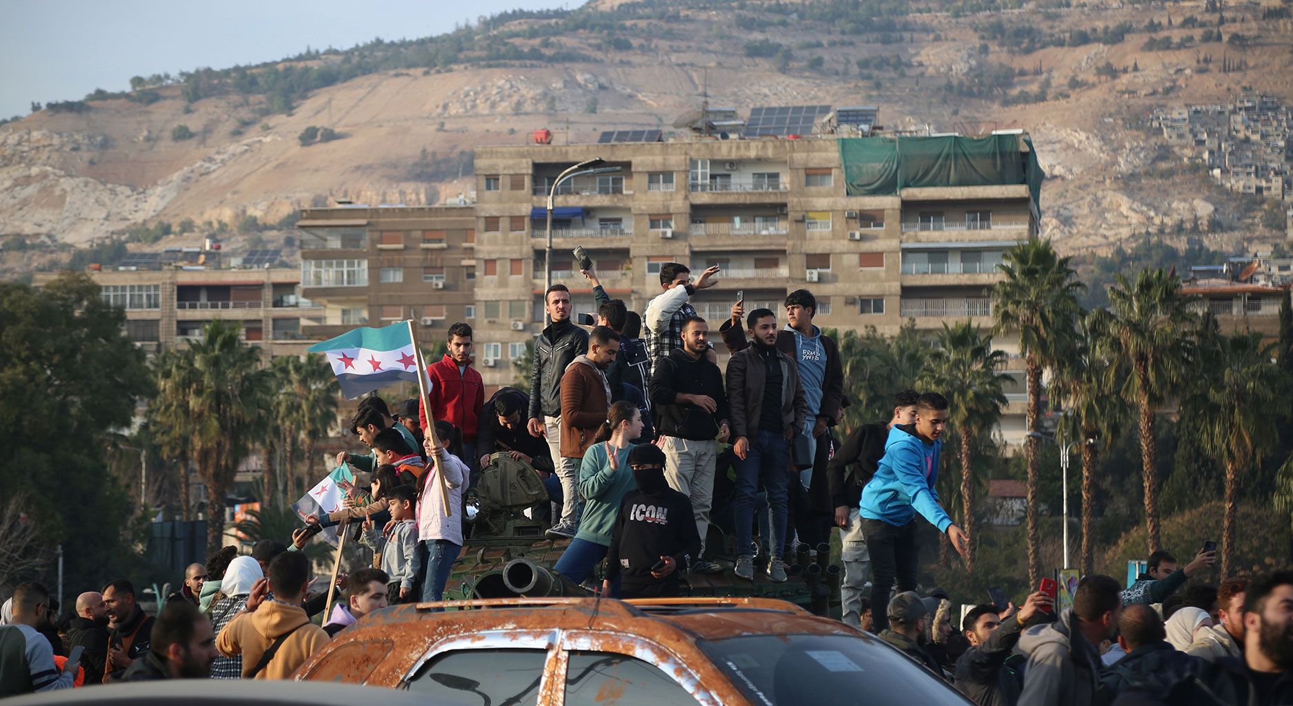 Menschenmenge mit Auto, auf dem viele Menschen stehen und eine Frau schwingt eine syrische Flagge. Im Hintergrund Palmen, Hochhäuser und ein Berg.