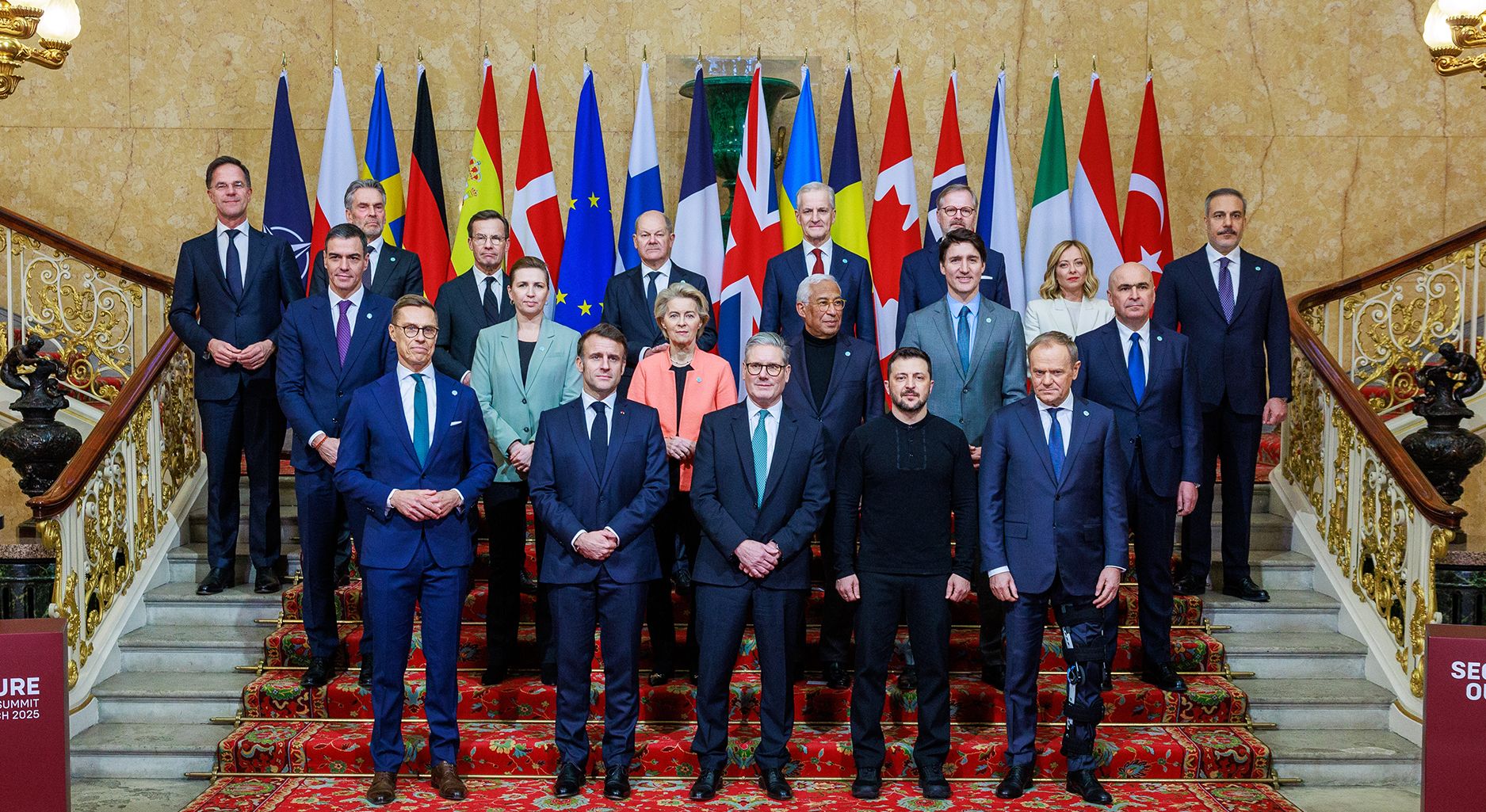 Gruppenfoto formell gekleideter Personen auf einer Treppe vor einer Reihe von Flaggen.