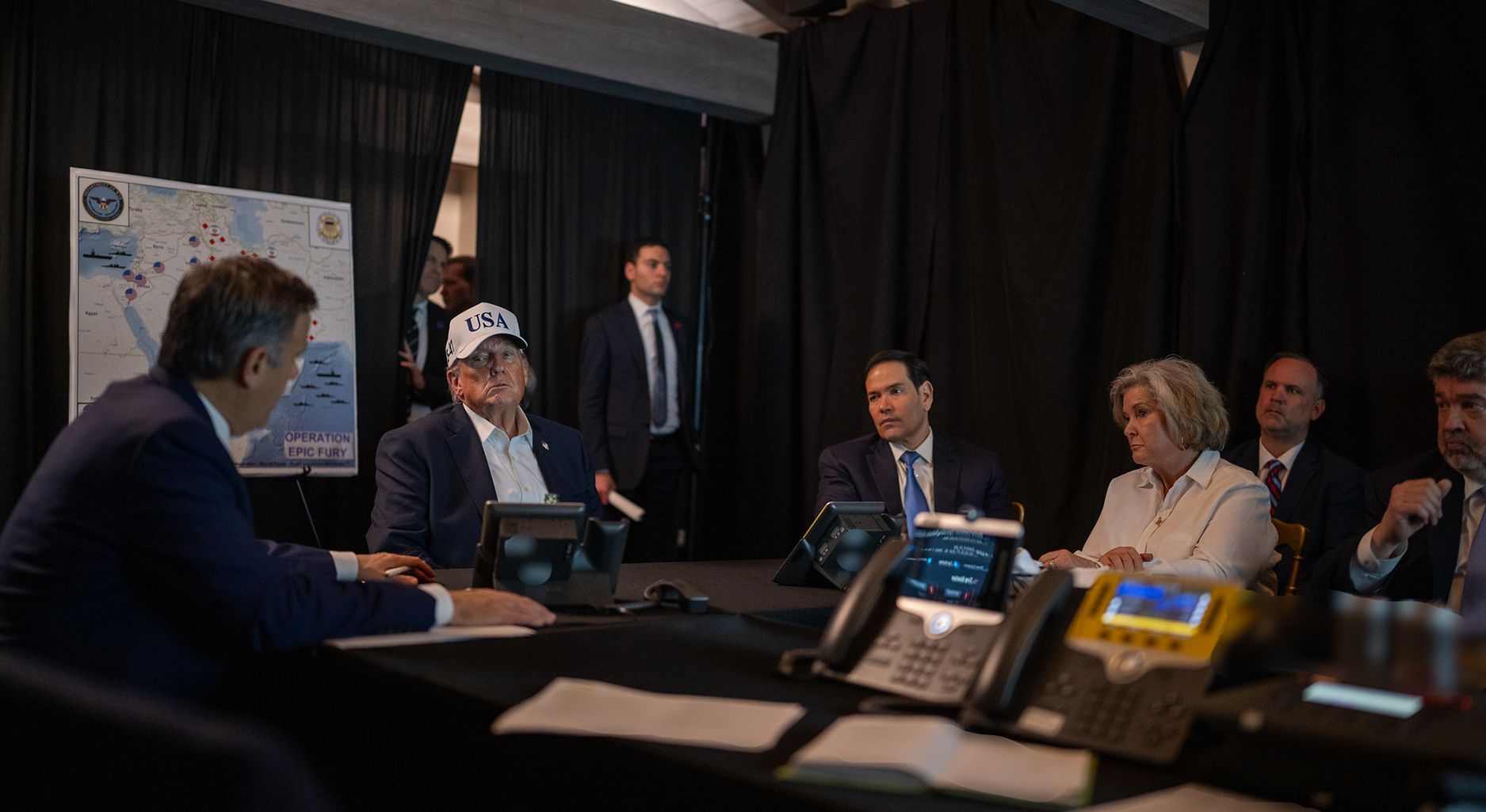 Donald Trump, Marco Rubio, Pete Hegseth and further staffers sitting in a room with curtains, telephones and a map of the Middle East on the wall.