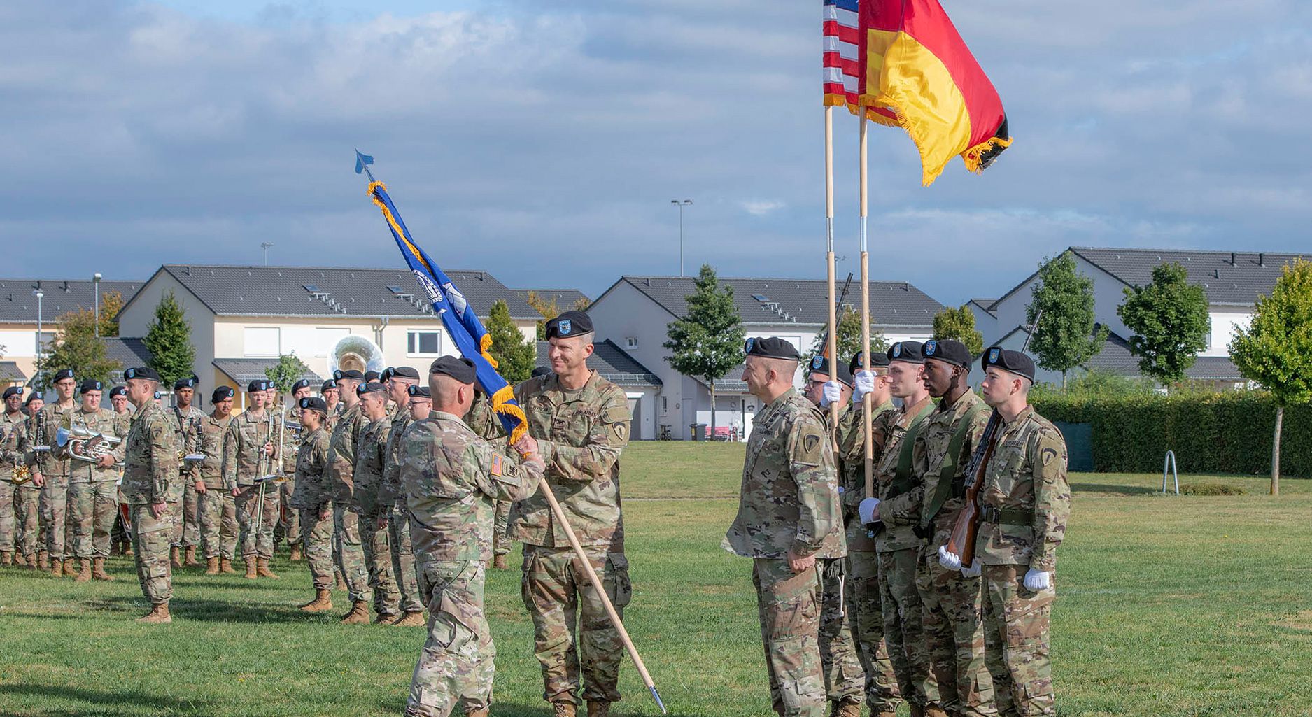 Ein Soldat überreicht lächelnd einem anderen Soldaten eine blaue, zusammengewickelte Fahne. Rechts von ihnen stehen sechs Soldat*innen, von denen zwei jeweils die deutsche und die US-amerikaische Fahne tragen. Sie befinden sich auf einer grünen Wiese, im Hintergrund sind links weitere Soldat*innen zu sehen. Dahinter befinden sich weiße Reihenhäuser.