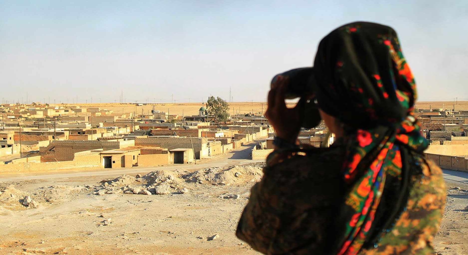 Person with black and red patterned headscarf seen from behind overlooking a settlement through binoculars.