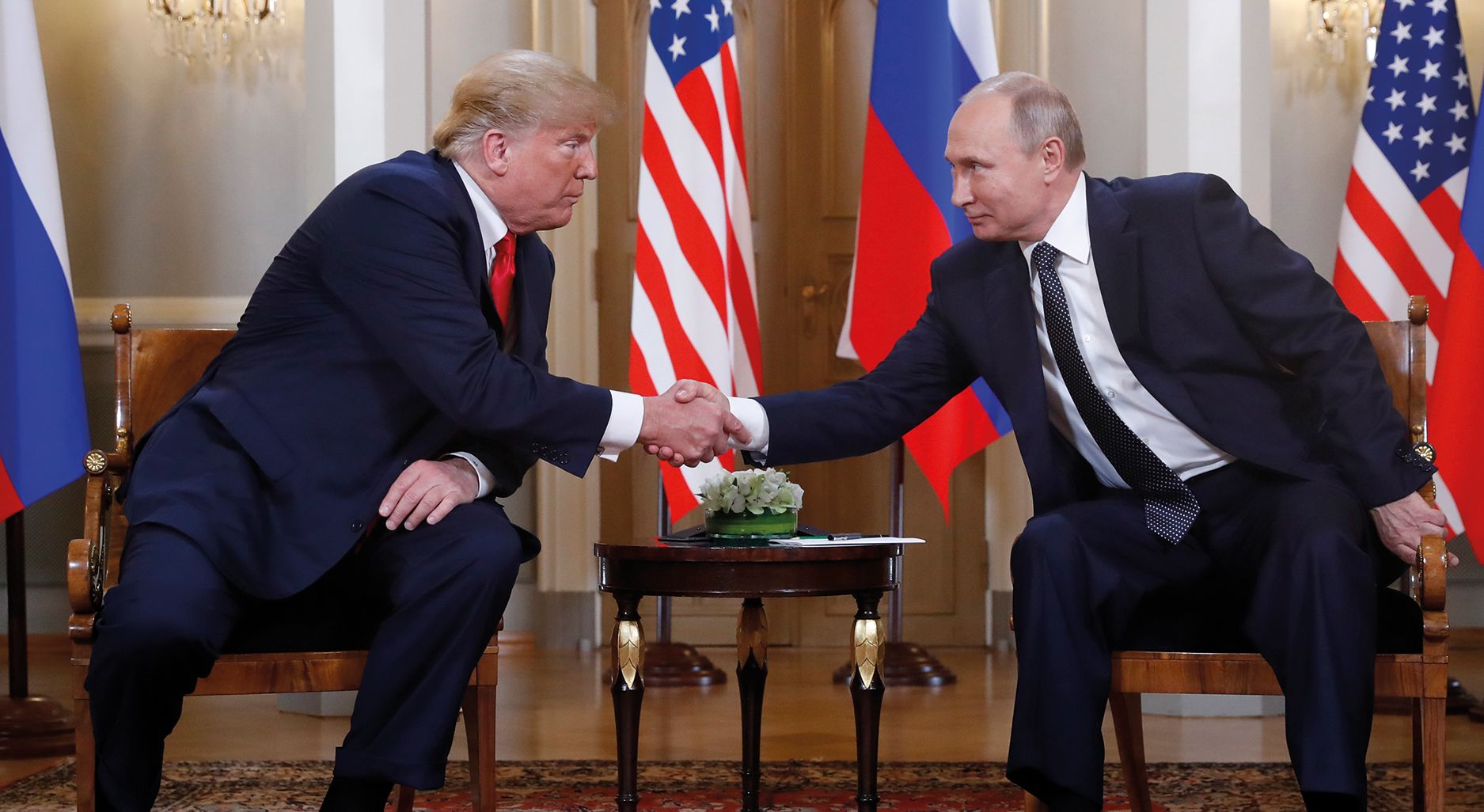 Two men sit on chairs and shake hands facing each other. American and Russian Flags in the background.