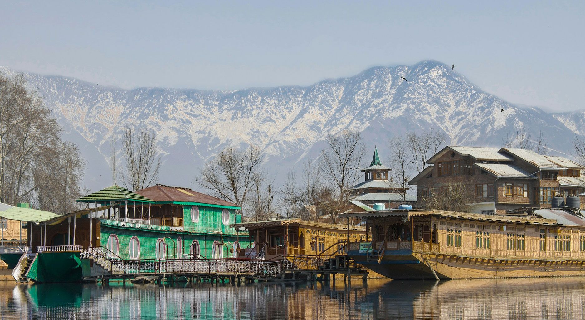 Photo of a green and a brown wooden house by a lake on which houseboats are floating. Snow-capped mountains rise up in the background.