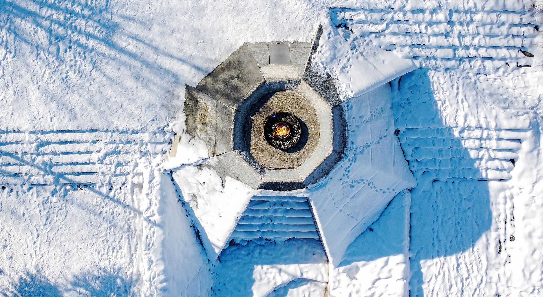 Aerial view of the “Eternal Flame Memorial.” It consists of a concrete platform with a fire bowl in the center where the fire burns. The memorial is partially covered with snow. There is also a lot of snow around the memorial.