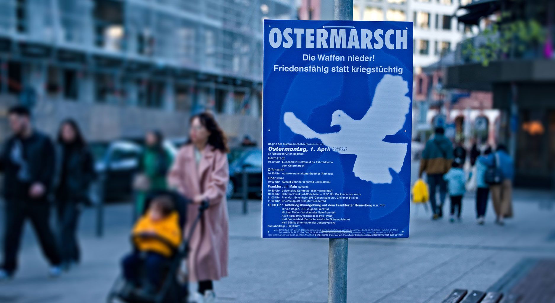 Laternenmast mit blau-weißem Plakat, auf dem eine Friedenstaube abgebildet ist und der Aufschrift "Ostermarsch. Die Waffen nieder! Friedensfähig statt kriegstüchtig". Im Hintergrund sind unscharf Menschen zu sehen.