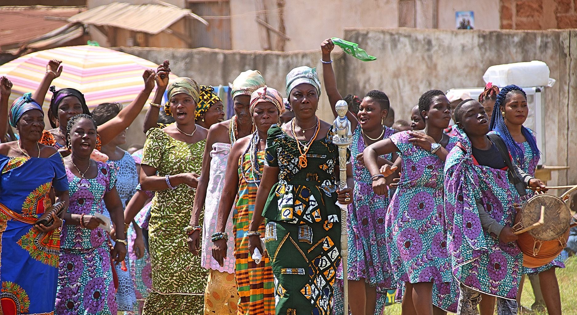 Group of colorfully dressed women walking. One woman is holding a drum, another a silver walking stick with a figure on top.
