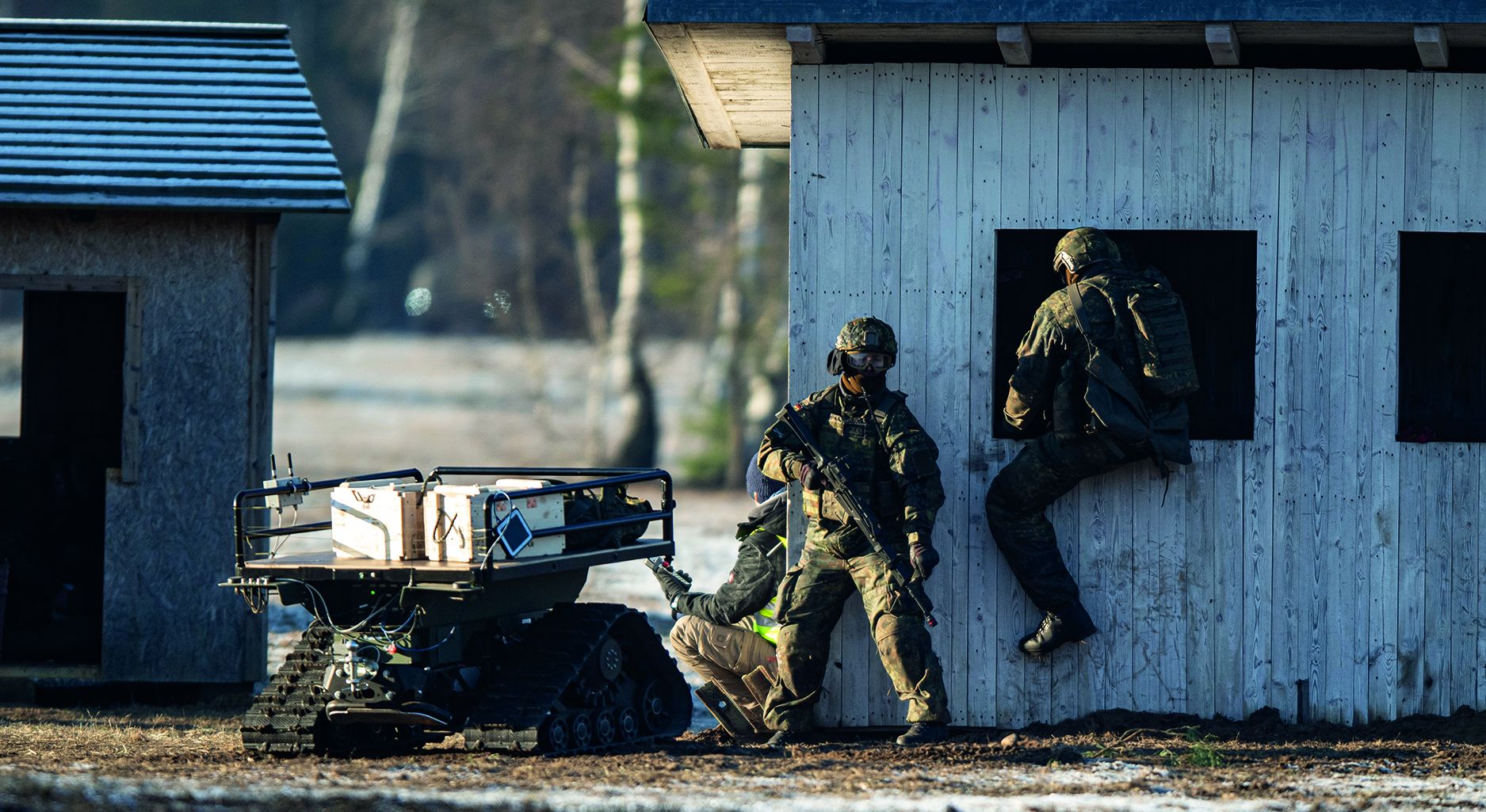 Image shows three soldiers, one of them climing into the window of a small wooden building, the other two holdling guns, next to a small vehicle with a flat top that holds several devices.