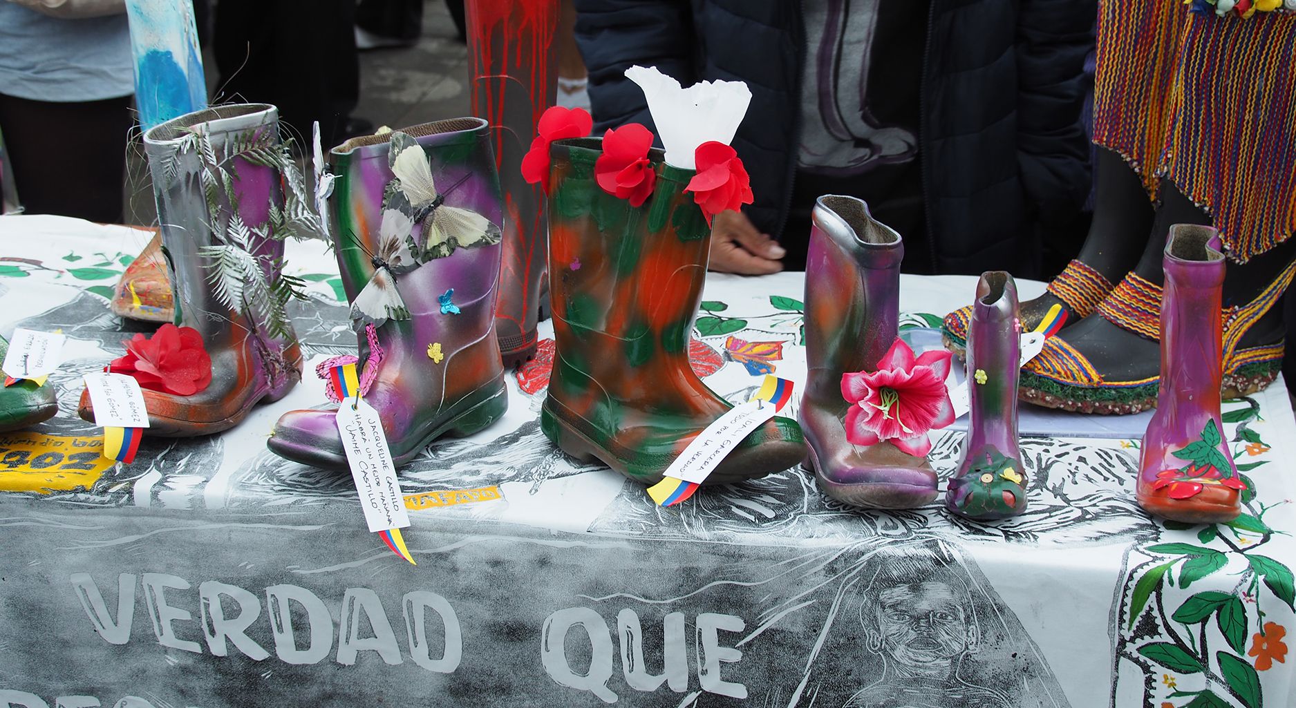 Boots decorated with various motifs such as flowers and butterflies are standing on a table outdoors, behind which you can see people wearing jackets.