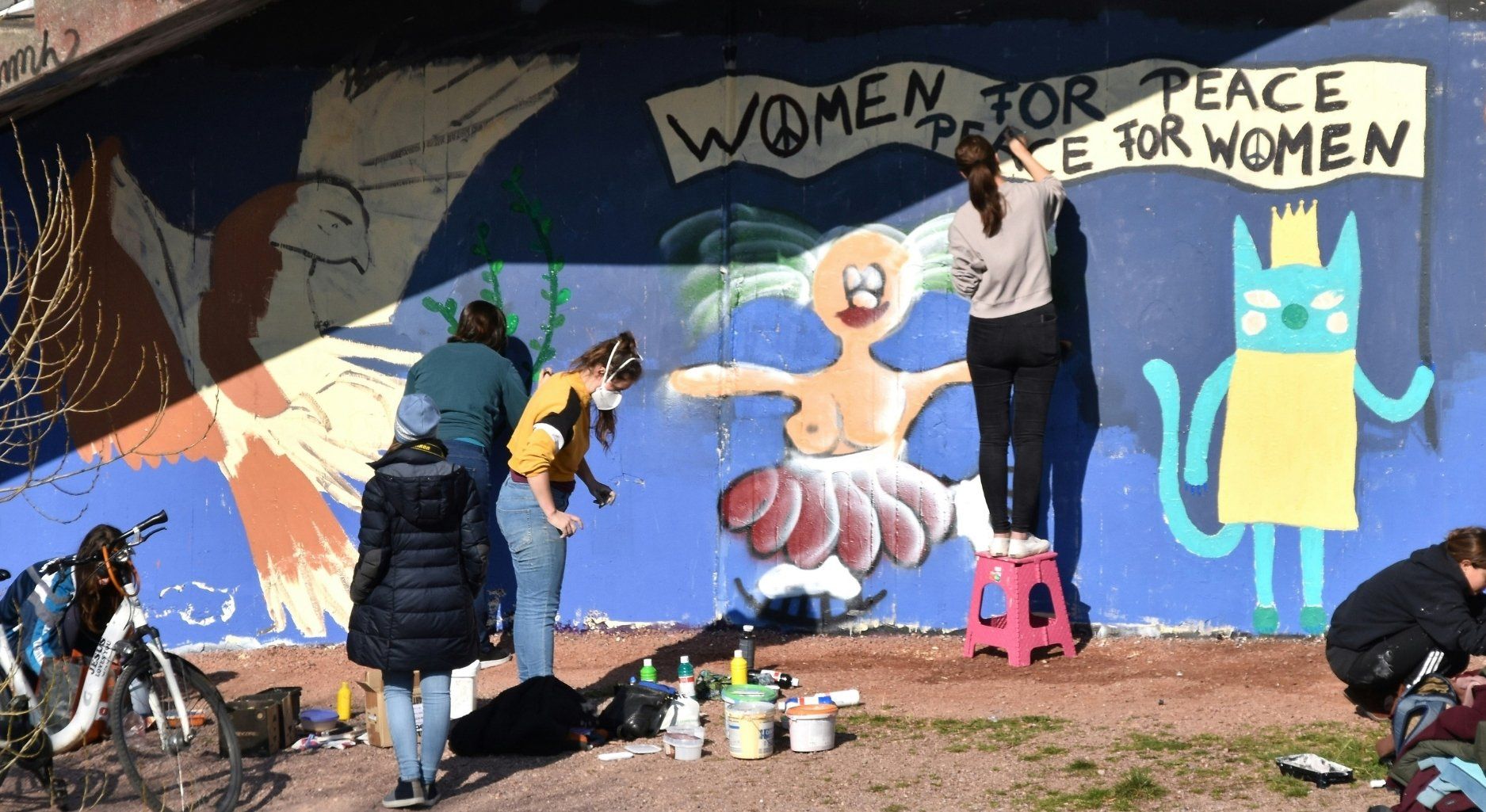 A group of people paint a mural with the words ‘Women for Peace - Peace for Women’ on the side of a building.
