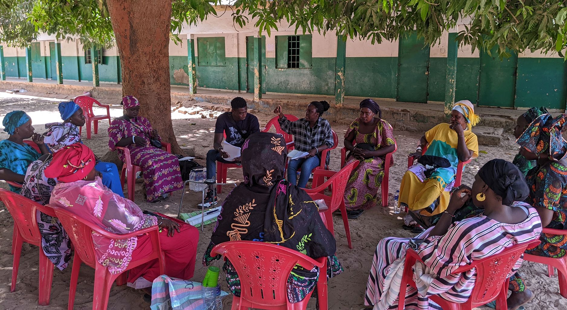 Group of people sitting in a circle on plastic chairs outside underneath a tree.