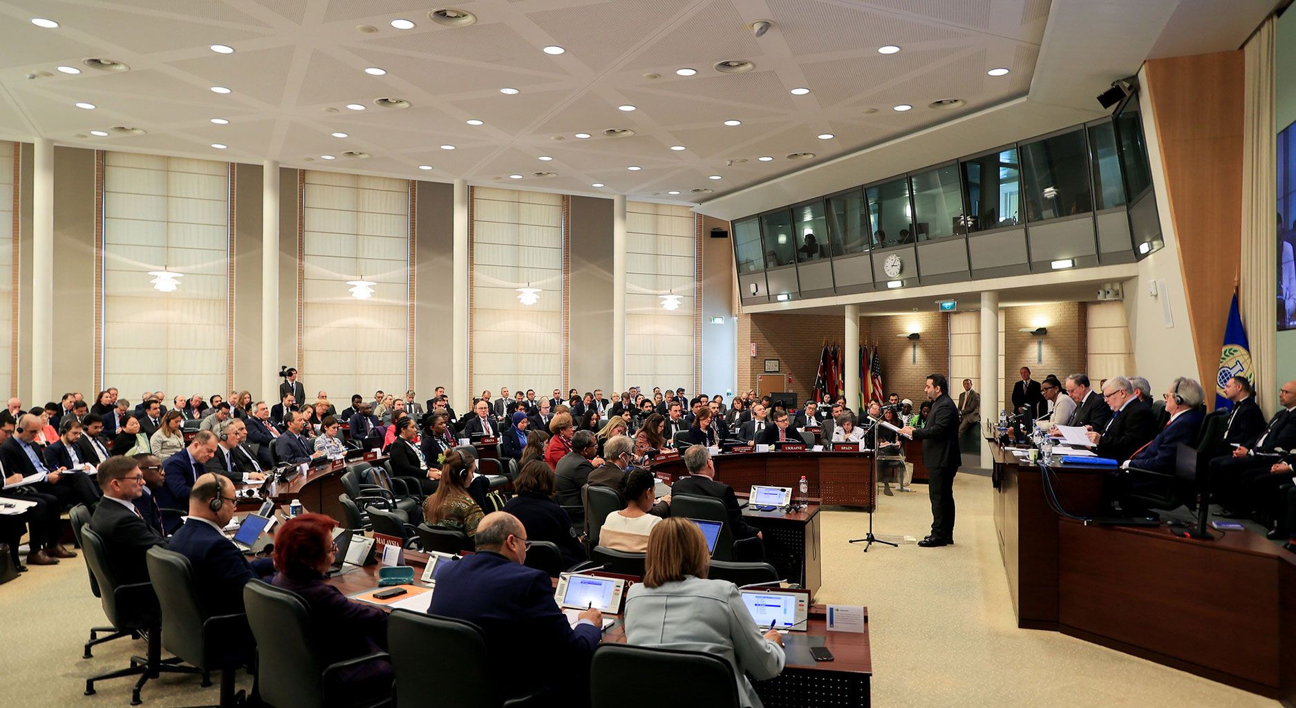 Room with semi-circular tables at which formally dressed people sit. A man in a suit is standing at the lectern, with a number of other people sitting behind him. The flag of the OPCW hangs on the wall.