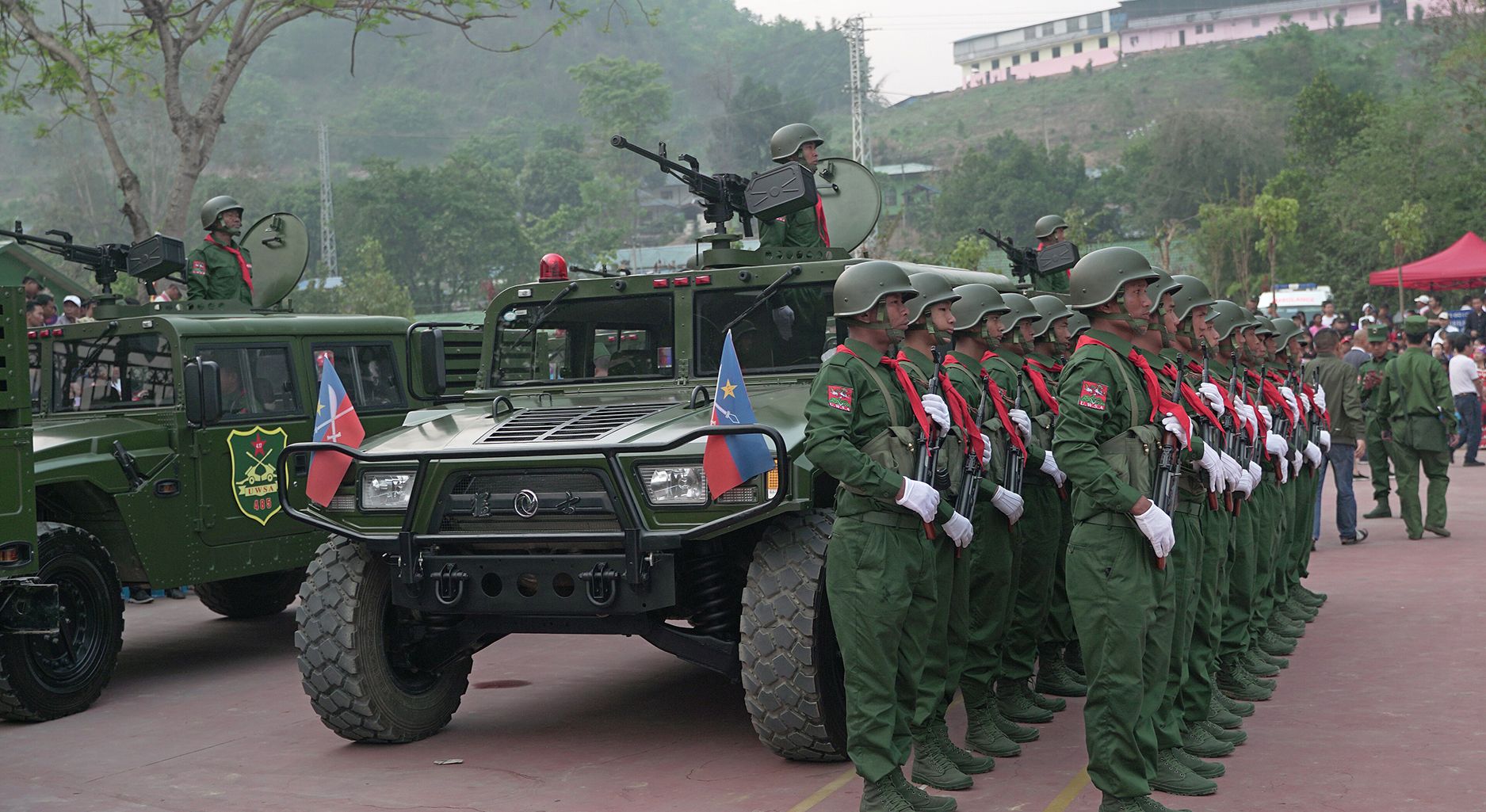 Two rows of soldiers in military uniform standing in front of military vehicles, with tree-covered hills in the background.