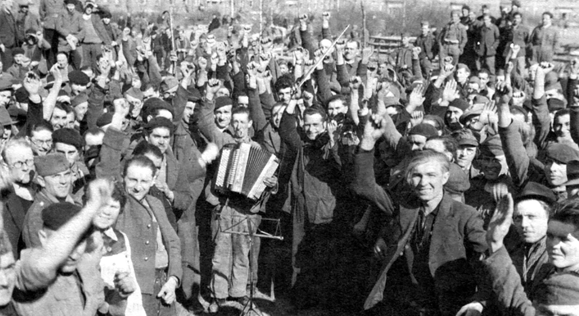 Schwarz-Weiß Foto. Eine große Gruppe Männer steht im Kreis. Sie lachen und reißen die Arme und Fäuste in den Himmel, ein Man trägt ein Akkordeon.
