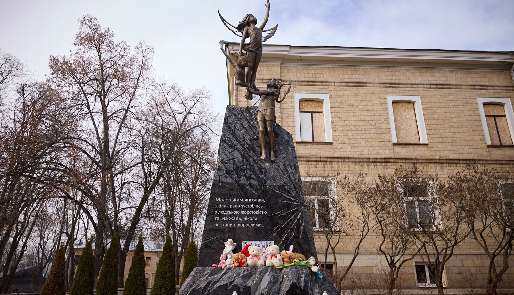 Im Bildvordergrund ist ein Denkmal: Das Denkmal besteht aus den Skulpturen eines Jungen und eines Mädchens in Form von Engeln mit verbrannten Flügeln, die über einen Granitblock fliegen, der eine Explosion darstellt. Im Hintergrund sind Bäume und ein Gebäude zu sehen.