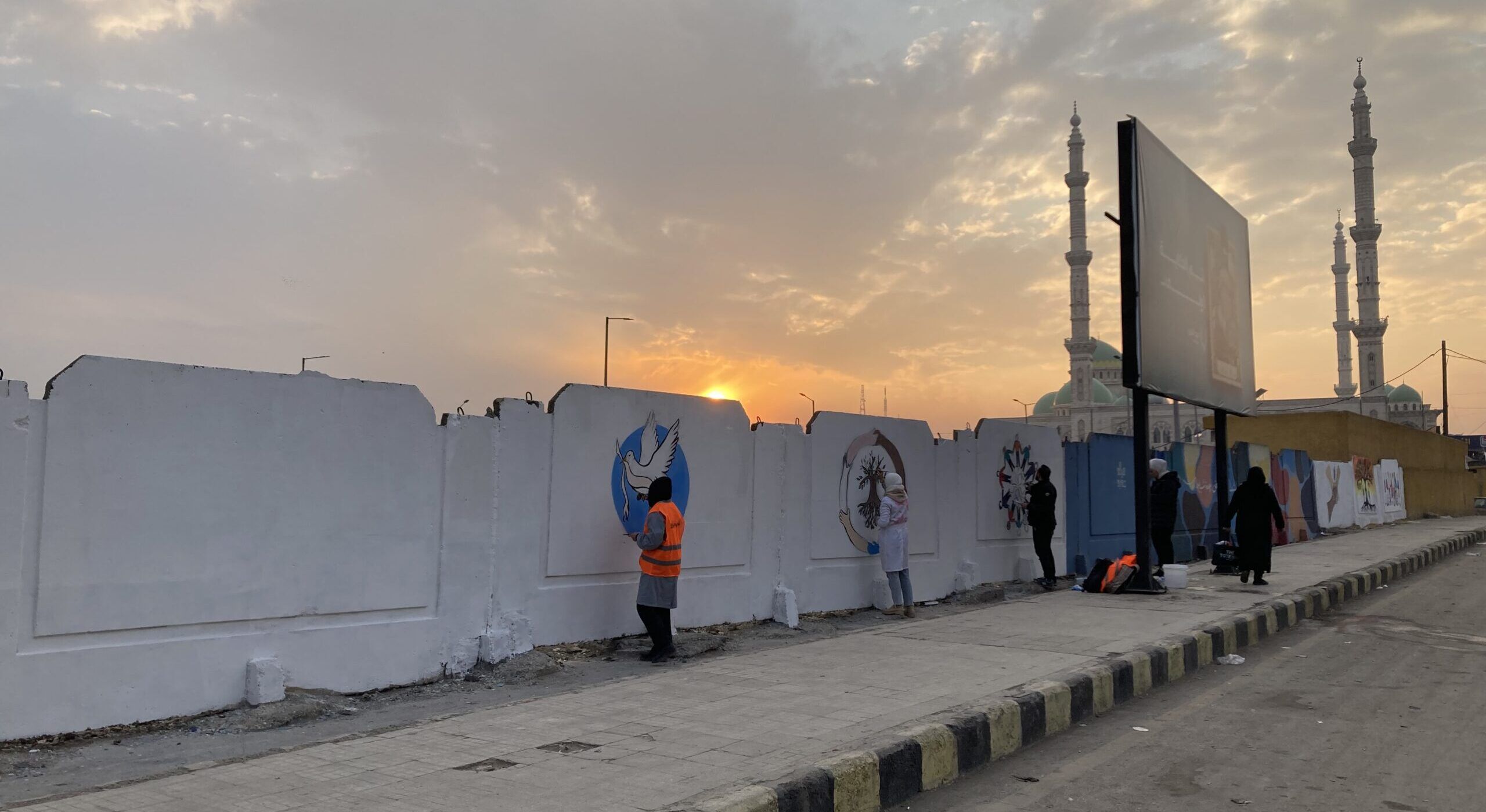 In the foreground, a woman wearing an orange vest is painting a dove of peace on a white wall. Other people nearby are creating murals, including another dove and abstract designs. The scene takes place on a wide street in Aleppo, with two minarets and a sunset visible in the background. The sky is cloudy and the lighting is subdued. A large dark sign stands to the right of the murals.