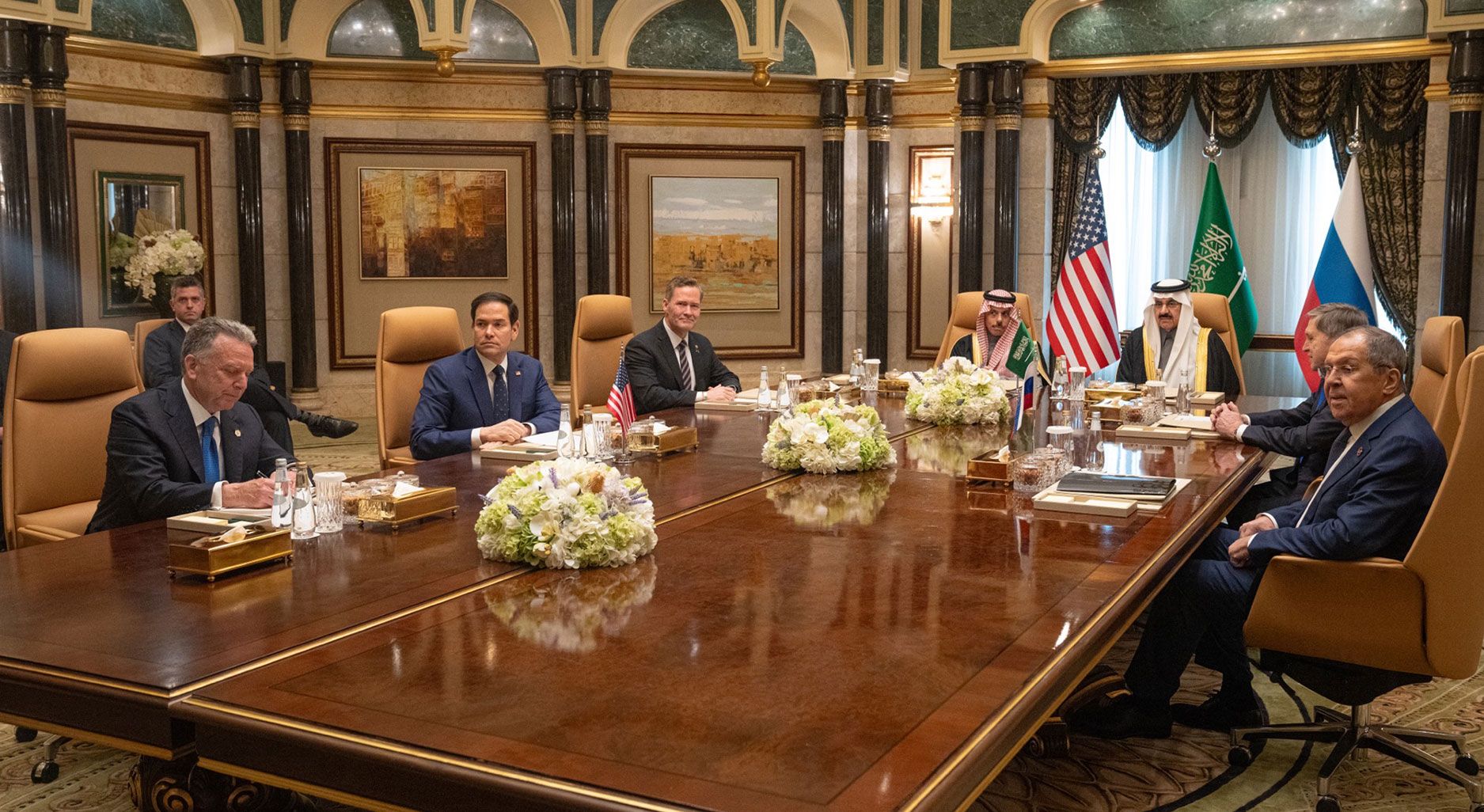 Photo einer Gruppe von Männern, die in einem prunkvollen Saal an einem Tisch sitzt. Dahinter sieht man die Flaggen von USA, Saudi-Arabien und Russland