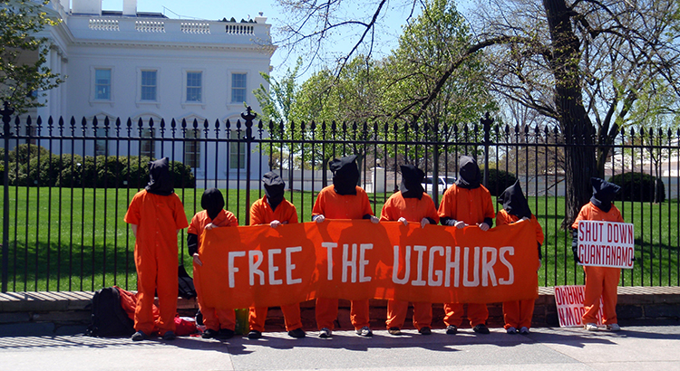 Protesters in front of the White House, dressed in red with black hoods over their heads holding a sign that reads: "Free the Uighurs". Another protester to the right holds a sign that reads "Shut down Guantanamo"