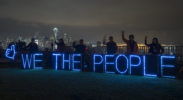 Nighttime scene with silhouettes of people holding glowing letters that spell out "We the people"