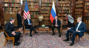 Image shows a meeting between Biden, Putin, Blinken and Lawrow as well as two translators. They are sitting in front of a wall of bookshelves and the US and Russian flags.