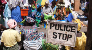 People sitting on a public square, in the foreground there is a sign saying "Police Stop".