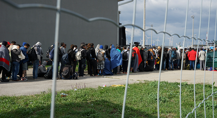 Eine Reihe syrischer Flüchtlinge überquert die Grenze zwischen Ungarn und Österreich auf dem Weg nach Deutschland. Durch einen Drahtzaun gesehen, im Hintergrund eine Windkraftanlage.