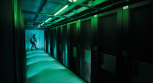 Grünlich beleuchteter Gang eines Serverraums, im Hintergrund geht eine Person vorbei