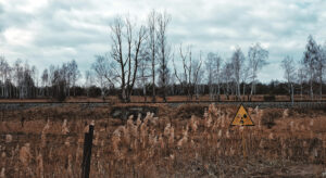 desolate landscape with trees and grey sky. A sign warns of nuclear contamination. With added logo “Socio-ecological Transformation Conflicts”