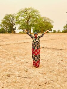Woman with colorful dress and headscarf standing in front of trees with her arms lifted